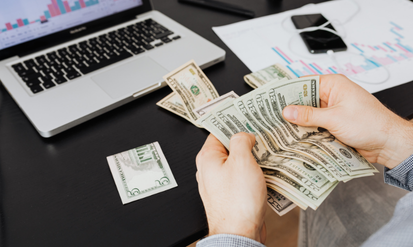 Person counting cash at a desk with financial charts and a laptop, representing private equity investment.